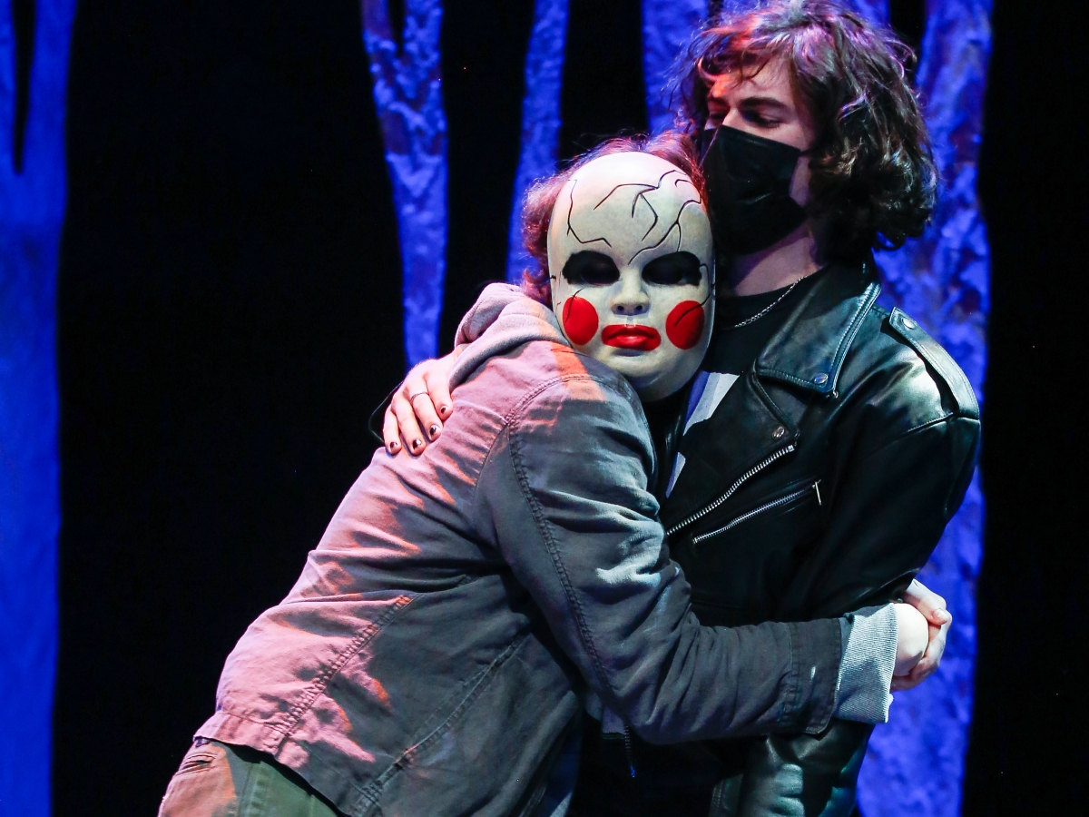 stage performance of hotspot. two people stand on stage awkwardly embracing against a background of blue velvet. the person on the right seems stiff, leaning slightly backwards in a black leather jacked and black surgical mask. the person on the left is clinging to the other, and has a creepy doll-like mask covering their face