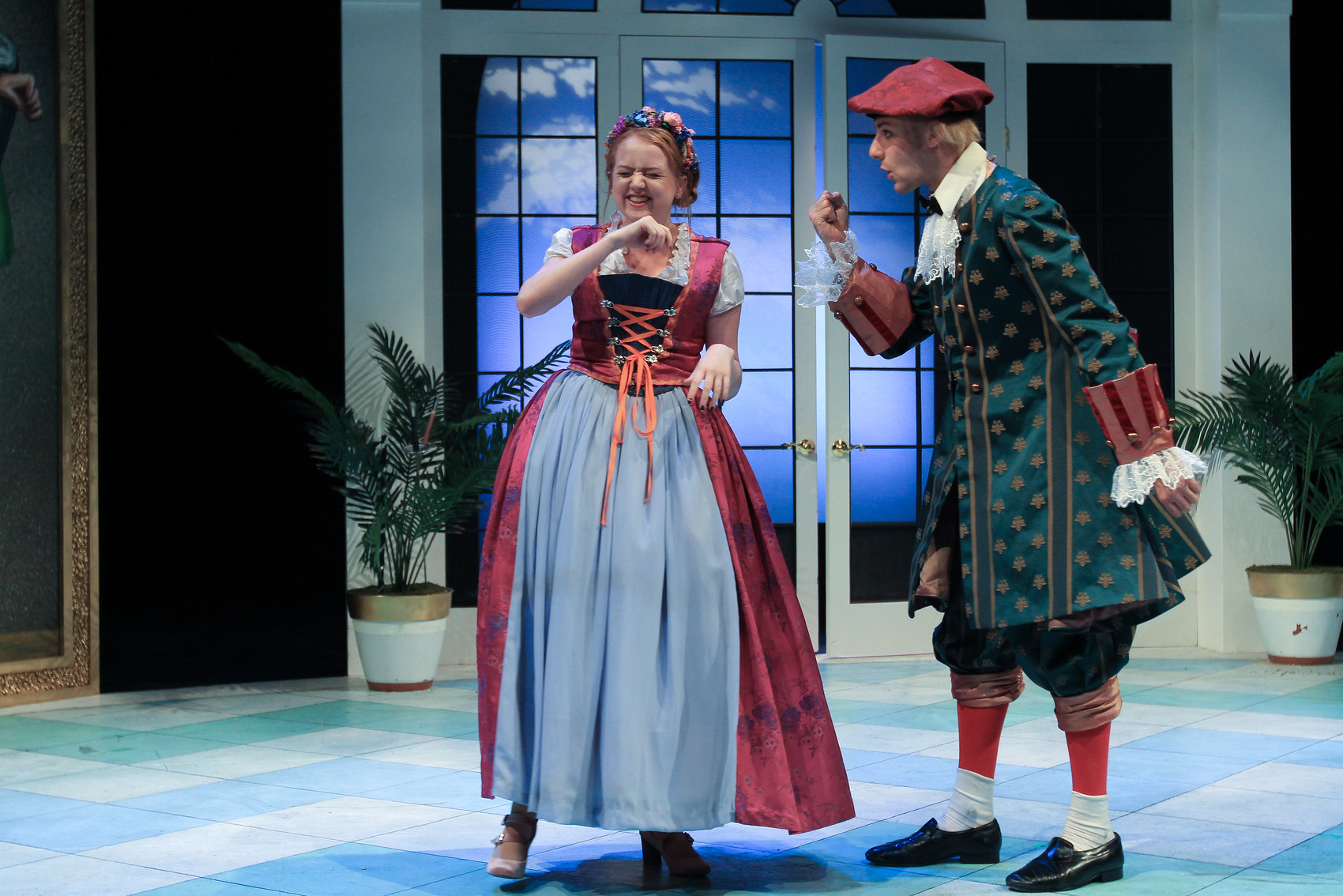 two student actors in period costumes stand on a stage of a checkered tiled floor and large windows, speaking at each other.