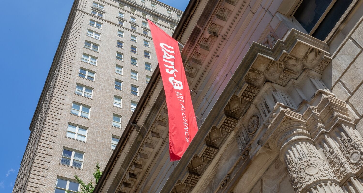 Image of the exterior of Art Alliance and another building against a blue sky, with a branded red UArts flag.