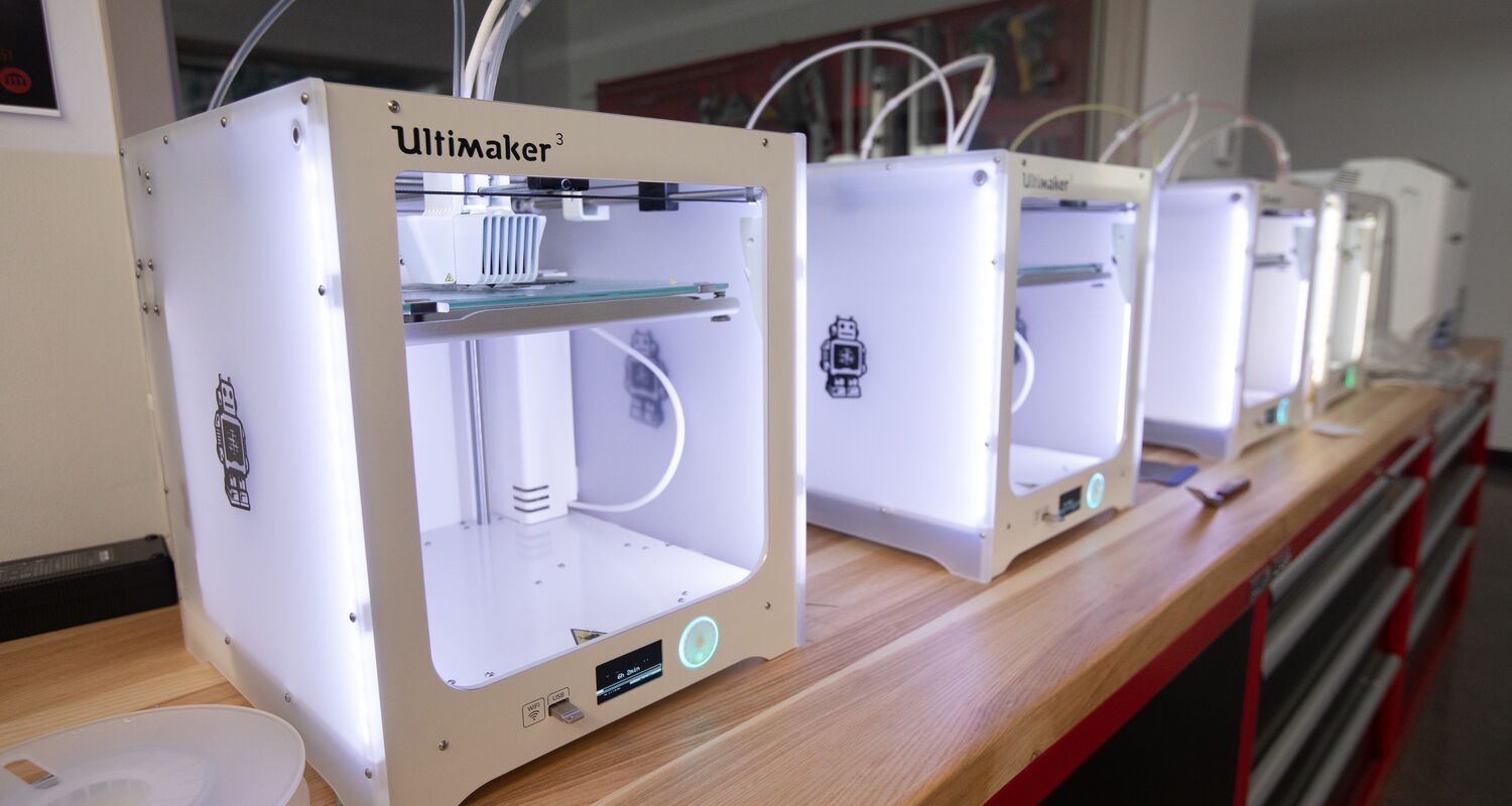 A row of 3D printing machines sitting on a workbench. The printers are white cubes with a cutout in the center.
