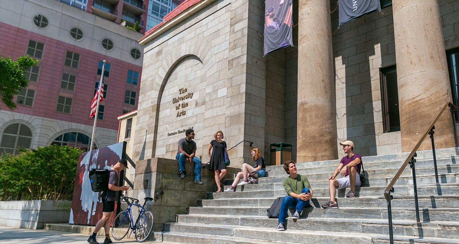 the steps of Hamilton Hall, an administrative building, with five students sitting on the steps and one student standing with a bicycle.