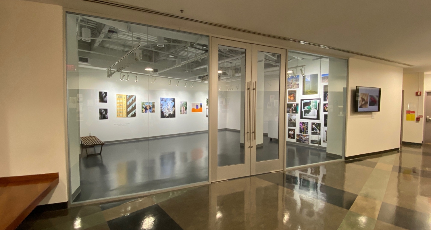 photo of the Photography Gallery from outside. the viewer is in a hall looking through glass walls and a large double glass door into a well light small white-walled gallery exhibition space with art photos displayed on the walls. 