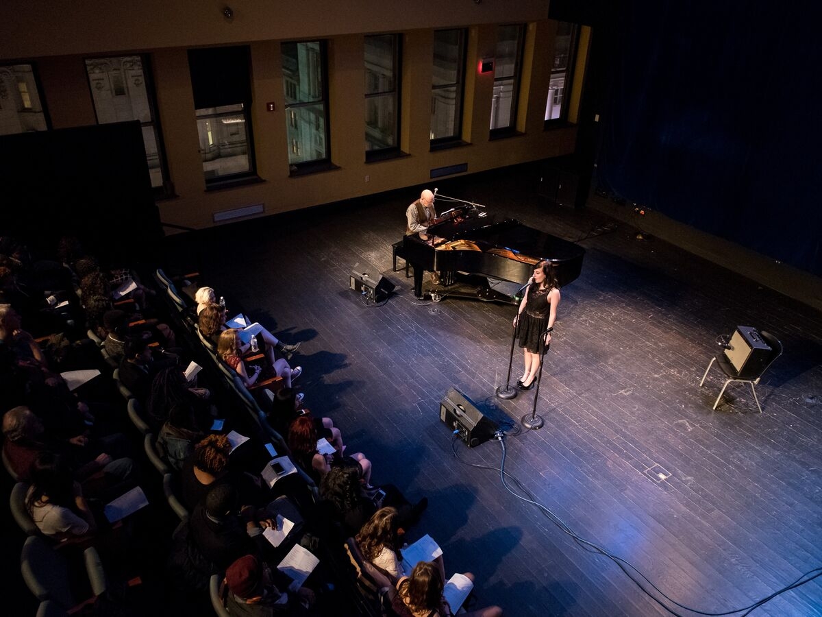 a vocal recital is seen from above. to the left are several rows of chairs with an audience facing towards the right of the image, where a single singer in a black dress sings into a microphone. a person in a brown suit vest plays on a grand piano towards the far wall, the windows of which reveal that the recital room is many floors up in a dense city. 
