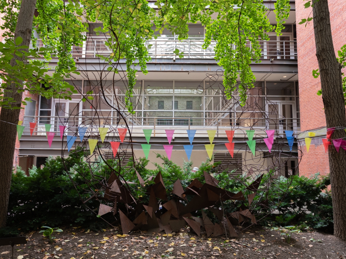 a view from within furness courtyard looking into hamilton hall's atrium. a rusted metal sculpture composed of waves of triangles is in the foreground, flanked by ginko trees whose bright green leaves hang from above. multi-colored pennants span between the two trees.
