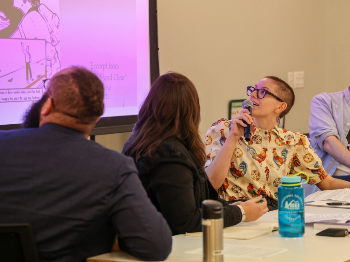 Bryn Ziegler MFA presenting at a panel. Ziegler is seated at a desk and looking over her right shoulder at a projection of a pink-hued artwork. Ziegler has huzzed hair and is wearing black horn-rimmed glasses and a button-up shirt featuring roosters. 