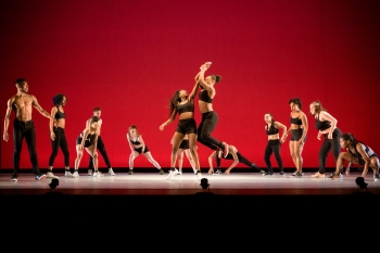 Dancers in front of a red backdrop are dressed in black tank tops and pants and shorts are in various stages of rising from and falling to the ground with two dancers jumping up in the middle 