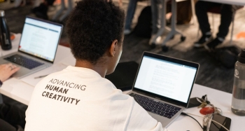 A student sitting at a laptop in class and wearing a white shirt with the words advancing human creativity on the back in black type