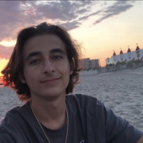 headshot of sean villa, shown in a dark grey t-shirt and a thing chain against a background of a sunset over a sandy beach.