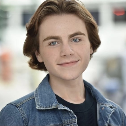 headshot of Kyle Edens. Kyle has medium length brown hair parted in the center and has a confident smile, wearing a blue denim jacket over a black shirt against ablurred out of focus background. 