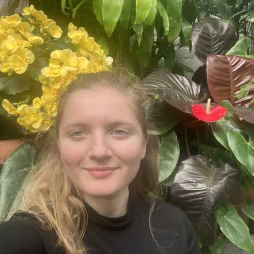 A young woman with blonde hair stands in front of tropical looking plants. She has a content look on her face and is looking just beyond the camera.