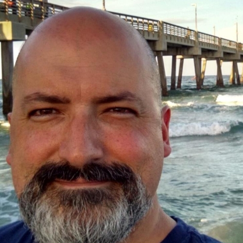 headshot of J Alex Cordaro. J is seen against a a background of a pier over choppy waves. J is bald and has a short grey bushy beard while wearing a blue short v neck t shirt. 