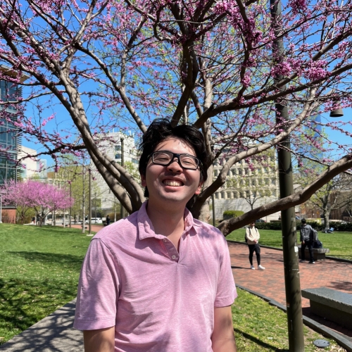 headshot of aki wakayama wearing a pink polo shirt and black glasses, smiling against a springtime park background with pink cherry blossoms bursting from trees in the background.