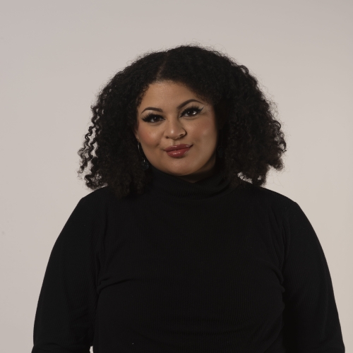 portrait of Z Murphy against a white background. Z has long curly dark hair and luscious lip stick on. she is wearing a black turtleneck tucked into flower-print red and black pants with her hands in the pockets.
