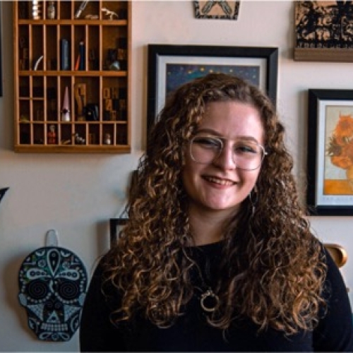 headshot of rachael miller against a background of artworks on a wall. rachael has long curly golden hair and round glasses