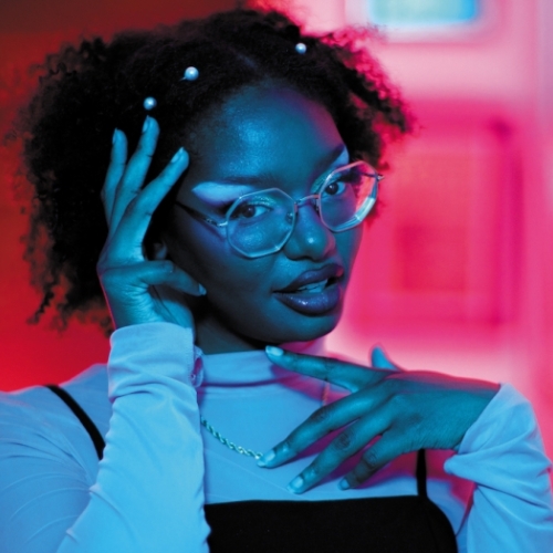 headshot of xenia matthews wearing a white turtleneck and glasses. matthews is bathed in blue light, while the background is a vivid fuchsia and red 