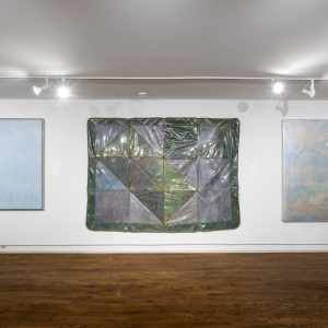 Invisible City Exhibition, Gallery E: a row of three large works on one long wall––one light colored geometric painting, one plastic encased quilt, one pale encaustic work