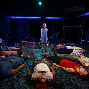 performance of buried. a person in a denim dress and red bandana stands in the middle of a room, surrounded by a circle of people lying flat on their backs. everyone is wearing face masks. the room is illuminated with dim blue lighting.