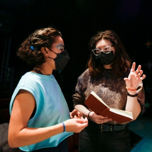 two people with longer curly hair pictured from about the waist up stand under a stage spotlight. the person on the right is further back and is holding a book in one hand with the other hand raise as if explaining something. the person on the left is wearing a blue sleeveless sweater and is gazing out of the frame.