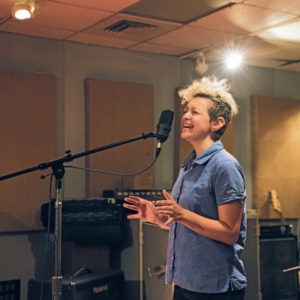 a person with a shock of bleached hear and a blue short sleeved collared shirt sings into a condenser mic in a treated recording studio with amps and recording equipment placed around the room.