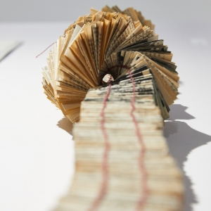 an artwork of complex folded paper bound into a long warping tunneling line, stretching from the viewer towards a curling spiral of pages. the paper appears to be pulled from book pages, as it is printed with text. it it bound with reddish thread. 