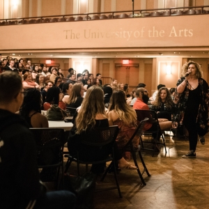 interior of Levitt Auditorium during new student orientation. The room is packed on the lower level of the auditorium, with all audience seating filled and additional folding chairs filled in the foreground. the upper balcony level is empty. all of the young people in the crowd are looking towards the right, where a standing person with long blond curly hair is walking towards the viewer and is speaking into a microphone. 