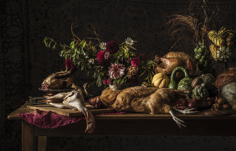 A dead, plucked chicken; dead chicken; and cooked chicken sit on a table with flowers