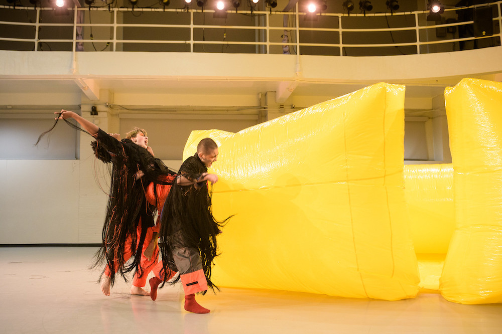 Two dancers in black and orange fringe costumes dance around a yellow sculpture