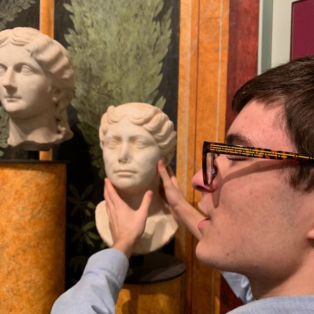 A visually impaired person in a blue shirt and tortoise shell glasses touching a sculptured marble bust