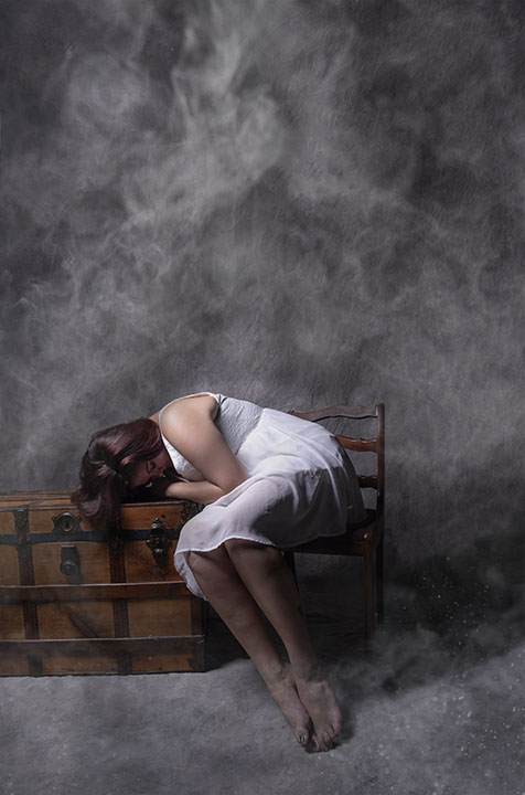 A female in a white dress sits in a chair slumped over a wooden trunk.