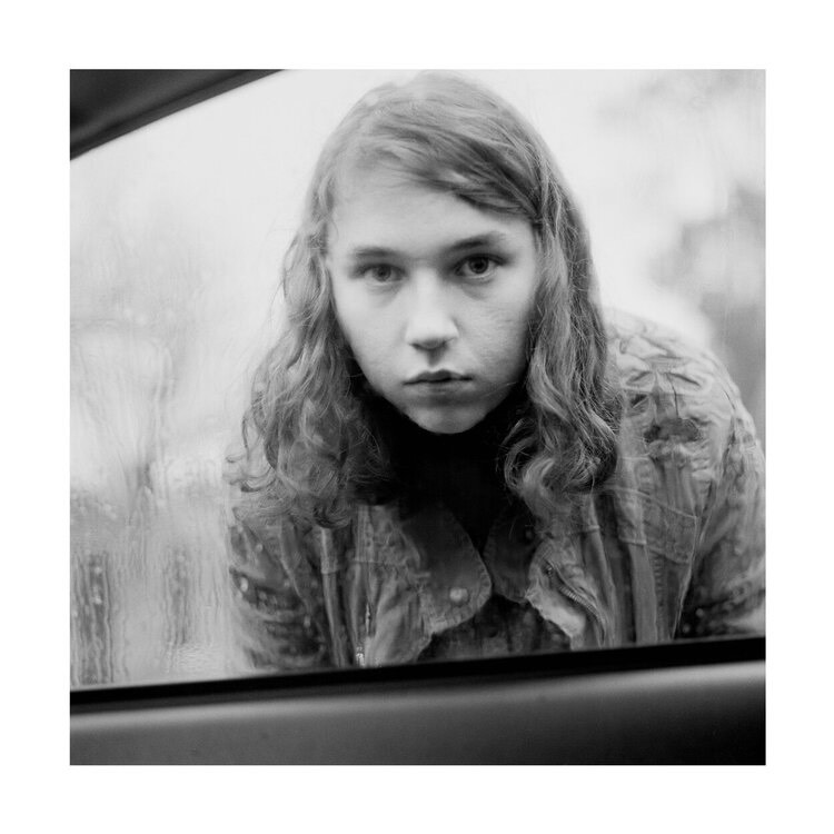 A photograph of a young person looking through a car window with rain