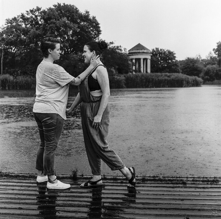 A photograph of two people standing close to eachother, one person has their hand on the others' neck. A pond and park are in the background. 