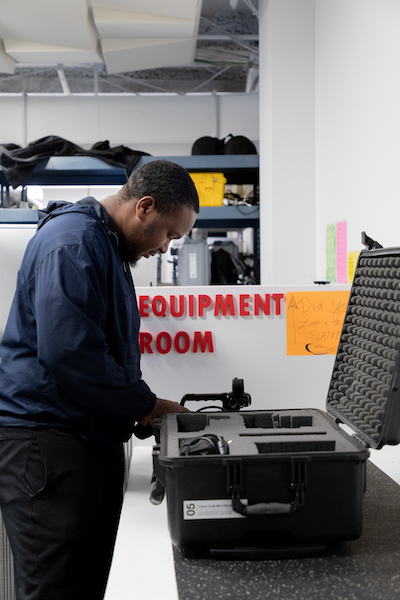 Student in Media Resources inspects a camera in a case. A sign that reads "Equipment Room" is in the background.