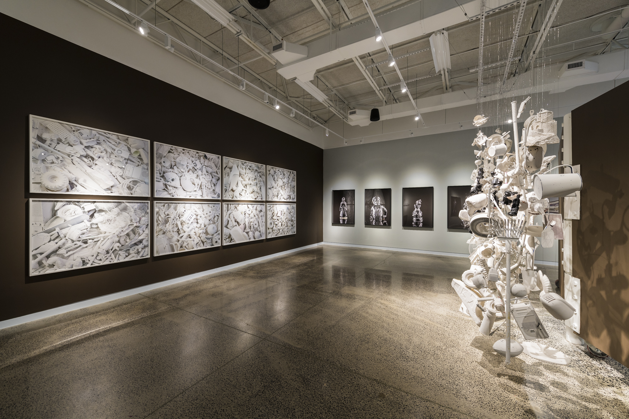 A gallery space with wooden floors and alternating wood and white walls with a six-panel abstract work hanging on one wall and a large white hanging sculpture made from household items hanging in the foreground 