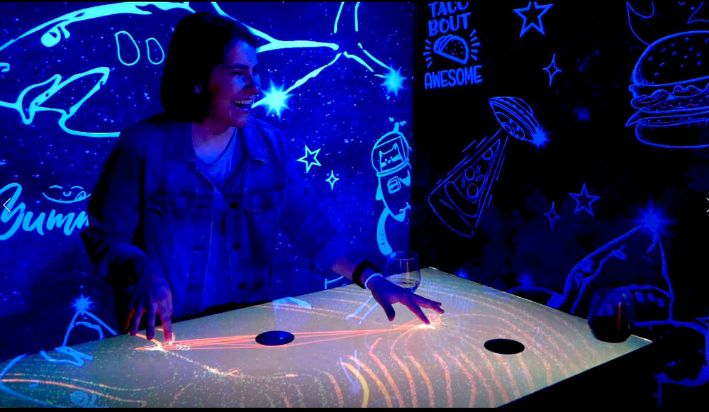 A dark room lit with iridescent/neon colored lights. A person standing at an interactive table appears to be drawing with light. 