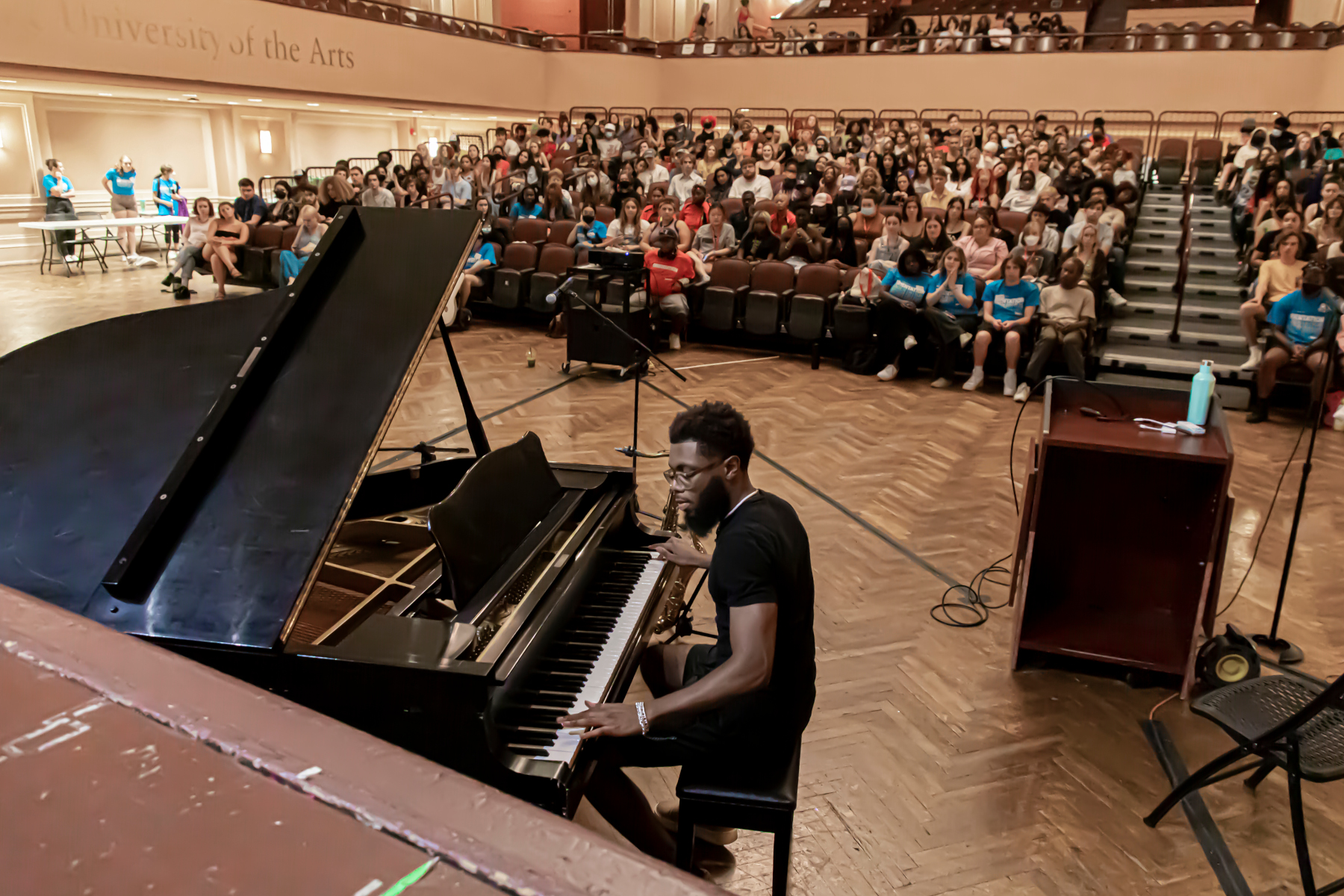 interior of levitt recital hall, seen from the stage. near the viewer is a grand piano with a person in a black t shirt and black glasses playing, seen in profile. the floor is a wooden chevron pattern. the raked seating area is packed with people. the seating area is ringed with a second floor balcony seating area