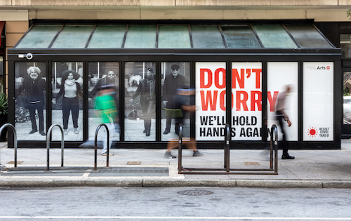 A window poster of people holding hands that reads "Don't Worry, We'll Hold Hands Again" from Resist Covid Take 6! by Carrie Mae Weems.