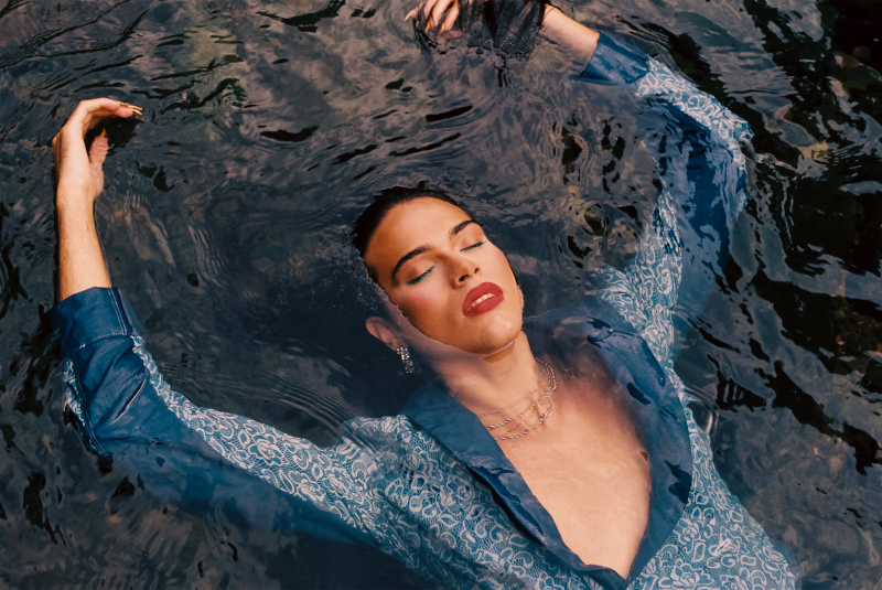 photograph by german vazquez depicting a person floating in rippling water face up with closed eyes, wearing sharp, bold makeup