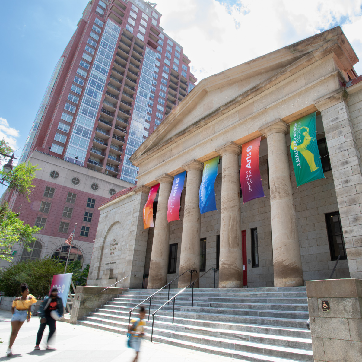exterior view of hamilton hall, seen at about 45 degree angle from facing the facade. a short flight of stairs leads up to the marble facade, where four pillars support an ornate carved roof element. five rectangular banners hang in the empty spaces around the pillars, with uarts logotypes displayed on beautiful color gradients. 