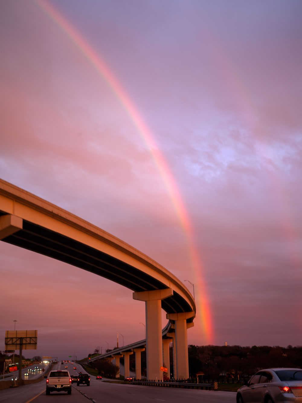A purple and pink sunset in the background of an elevated roadway with a rainbow extending almost vertically to the top of the image