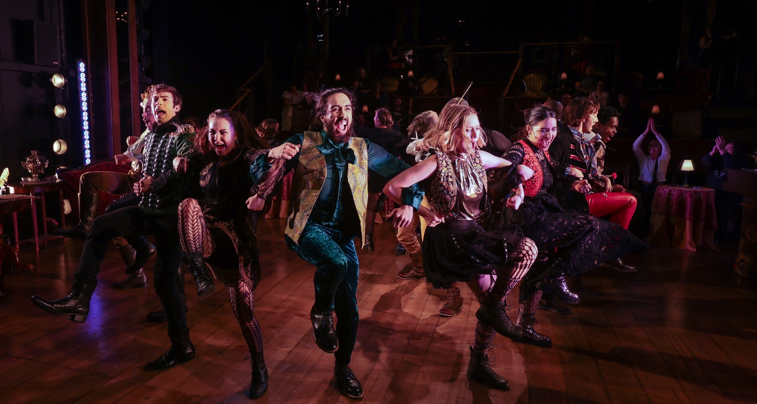 A group of students dancing with their arms linked. The energy is high, everyone is smiling. The floor is brown wood, and the costumes are a mix of colors including green, red, gold, and black. The image is from the Brind School production of Natasha, Pierre & the Great Comet of 1812 By Dave Malloy.