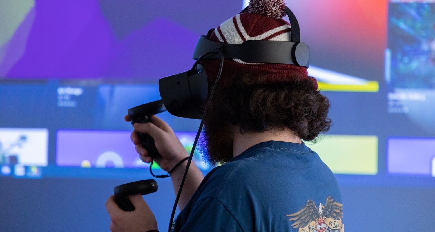 A student with thick curly hair and a beard wears a beanie and a VR headset. He is holding VR controllers, facing away from the viewer. A projected interface is visible on a screen in front of him.