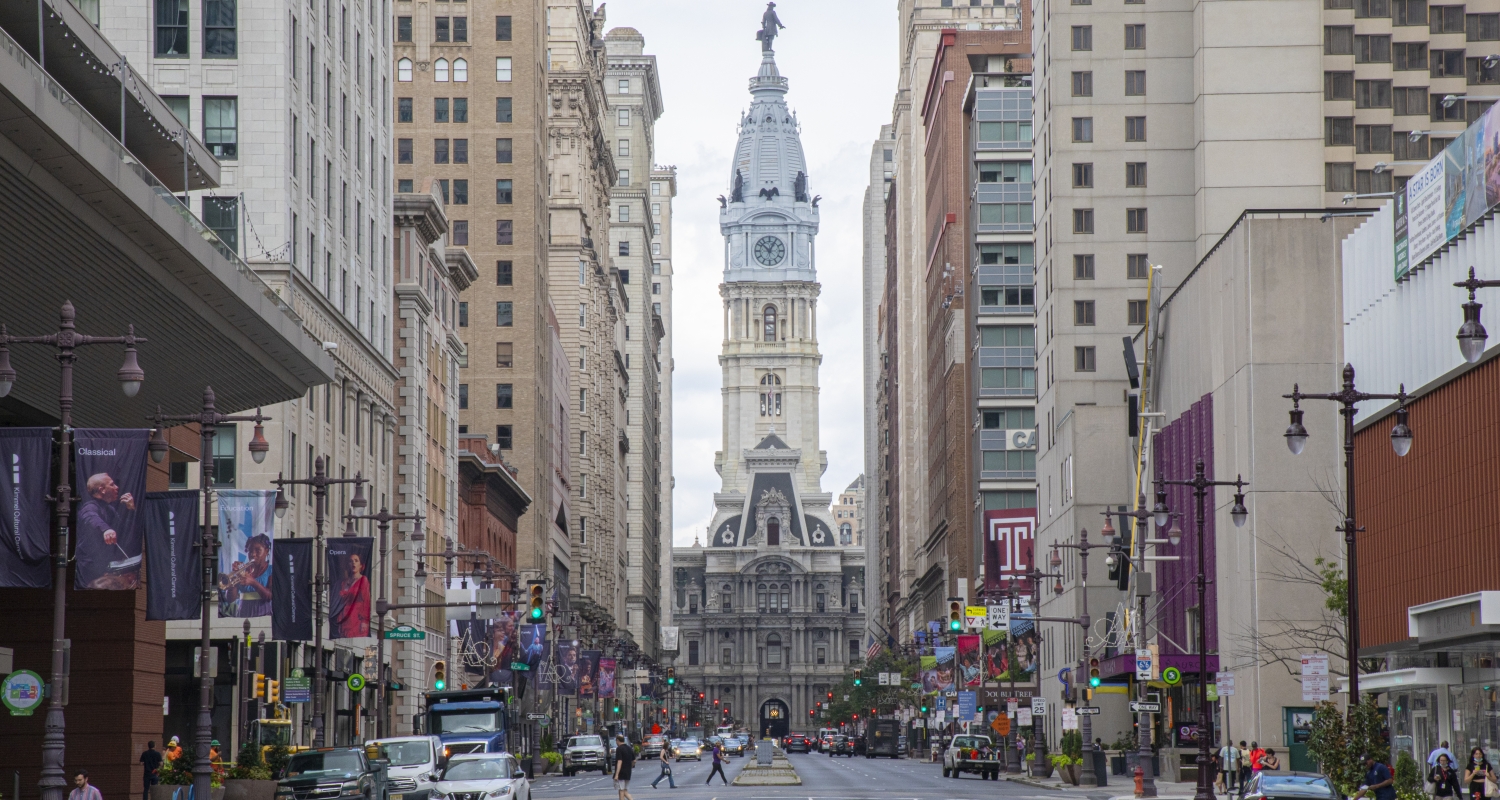 A view down the Avenue of the Arts, a section of Broad Street in Center City Philadelphia, that highlights the iconic City Hall building situated by several tall buildings on either side. 