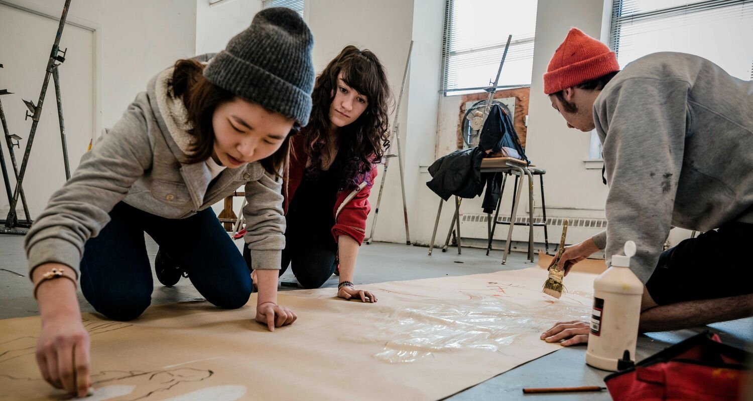Three students kneeling on a studio floor, working on a large drawing on brown paper. 