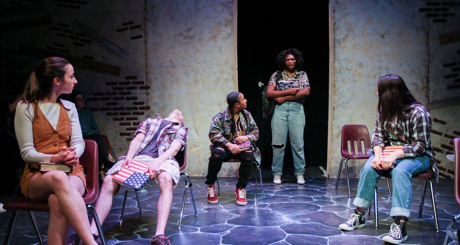 Students on stage for an Equinox performance ("Boxed", 2019). A student stands in the background, center stage, with her arms crossed. The other students are looking over their shoulders at her: a student seated at the far left looks over her shoulder wearing a tan dress; directly behind her a student is leaning back in his chair with a book or laptop in his hands. A third student on the left, but closer to the center, is wearing a multi-colored sweater; the last student is on the right with a plaid shirt.