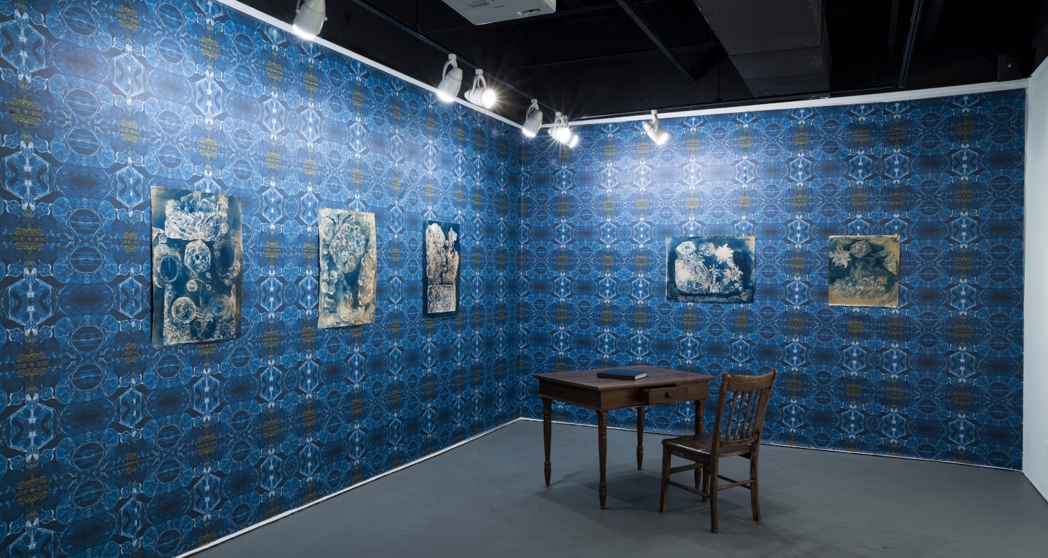interior of a gallery, with a grey floor and a small, ornate wooden desk and chair towards the back between the walls. The walls are blue with a complex pattern similar to ornate ceramic tiles. there are bluish images hung on the walls, illuminate with bright white lights from above. 