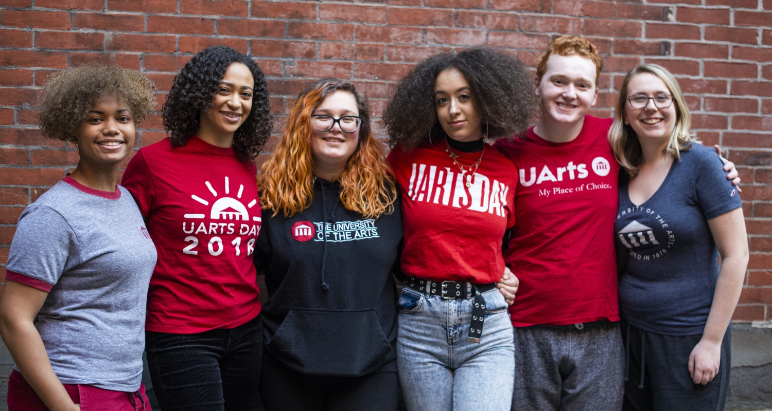 UArts students wearing a variety of UArts T-shirts stand against a brick wall with arms around each other
