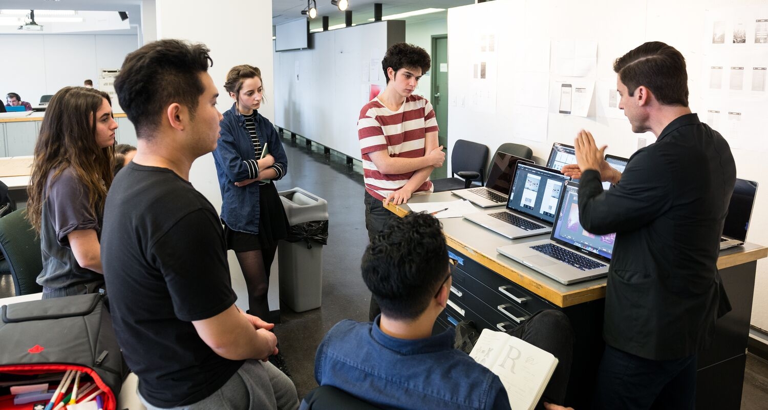 Students, both standing and seated, in a graphic design classroom gather around several laptops on drafting table. An instructor on the right is gesturing and the students have engaged and thoughtful facial expressions.