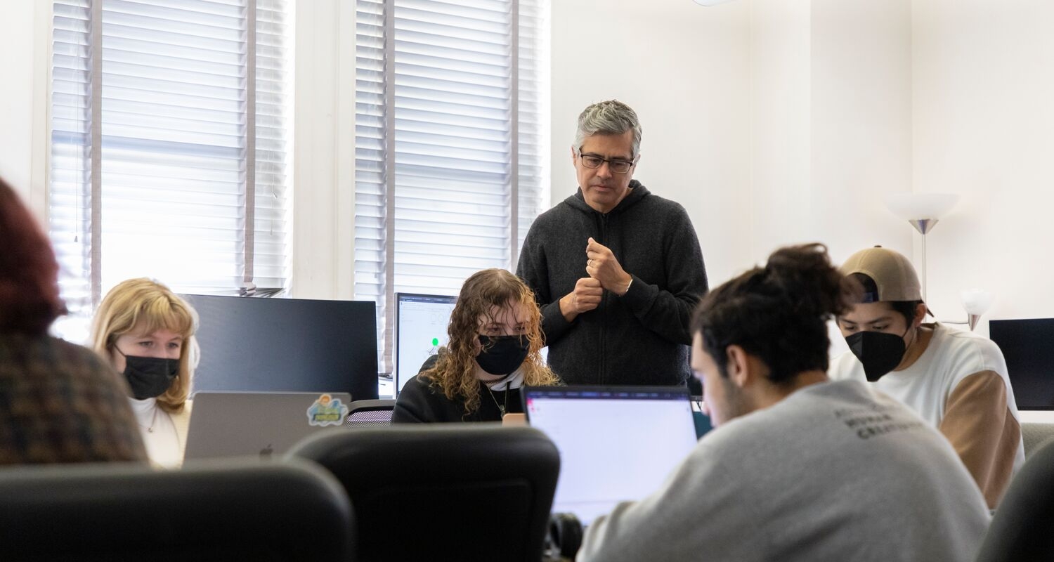 Juan Parada, Interaction Design program director, stands in the middle of a light-filled classroom with several students who are sitting at laptops at a table in front of him.