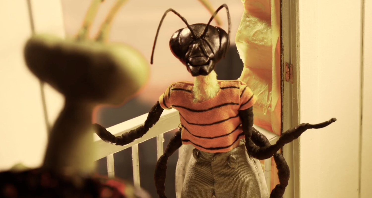 a still from stop motion film Lemoncholy still by Maya Pirulli and Mariluz Rodriguez depicting a bee doll wearing an orange striped shirt and pants facing forward talking to a praying mantis seen from behind in an over the shoulder shot. 