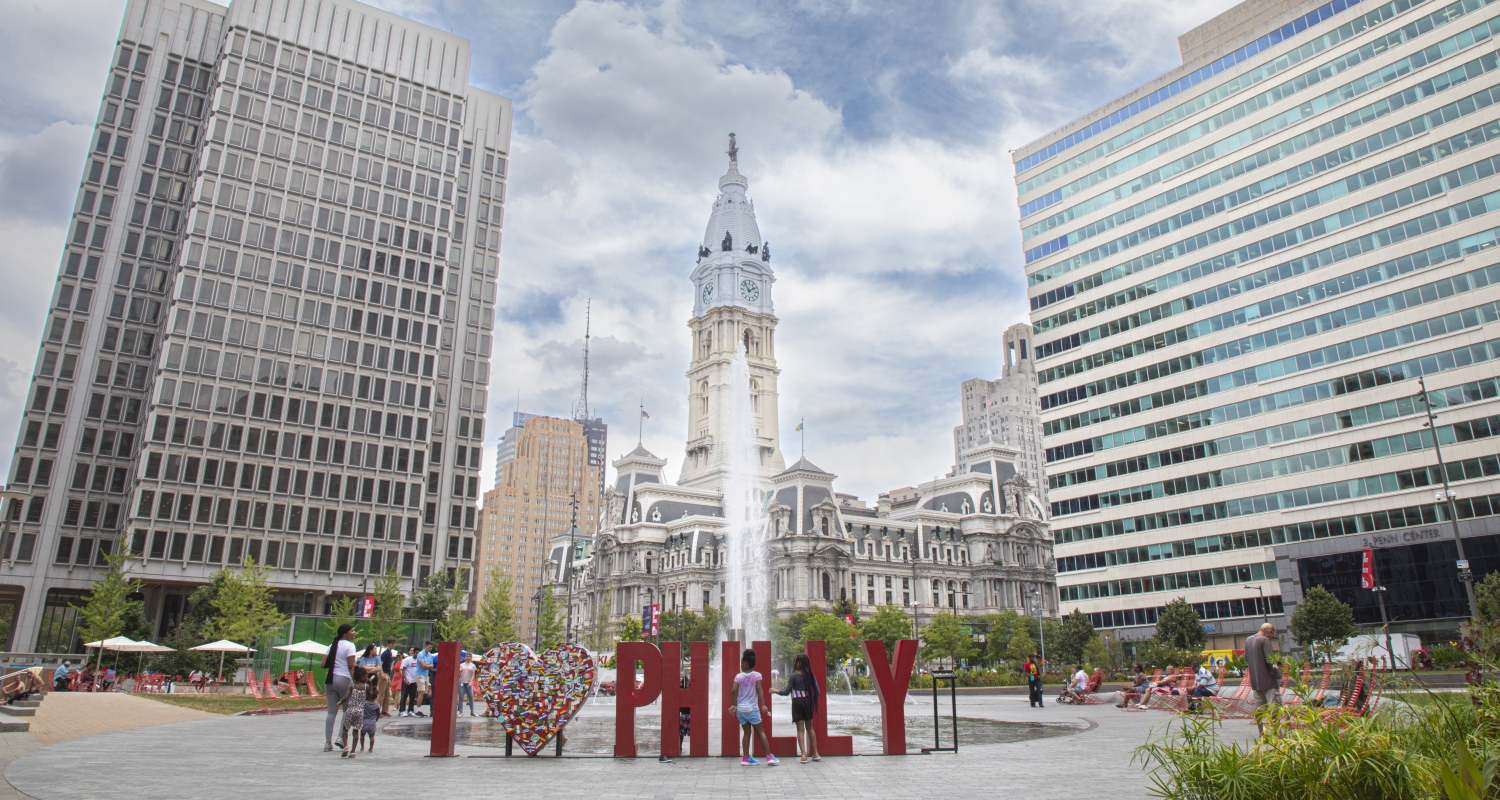 Letters that spell "I [heart] Philly" are central to this photograph of a dynamic view of Center City Philadelphia. People are gathered by the letters giving a sense of scale. Two towering corporate or residential buildings flank the iconic City Hall building. Water splashing vertically up from a fountain is also central to the scene.